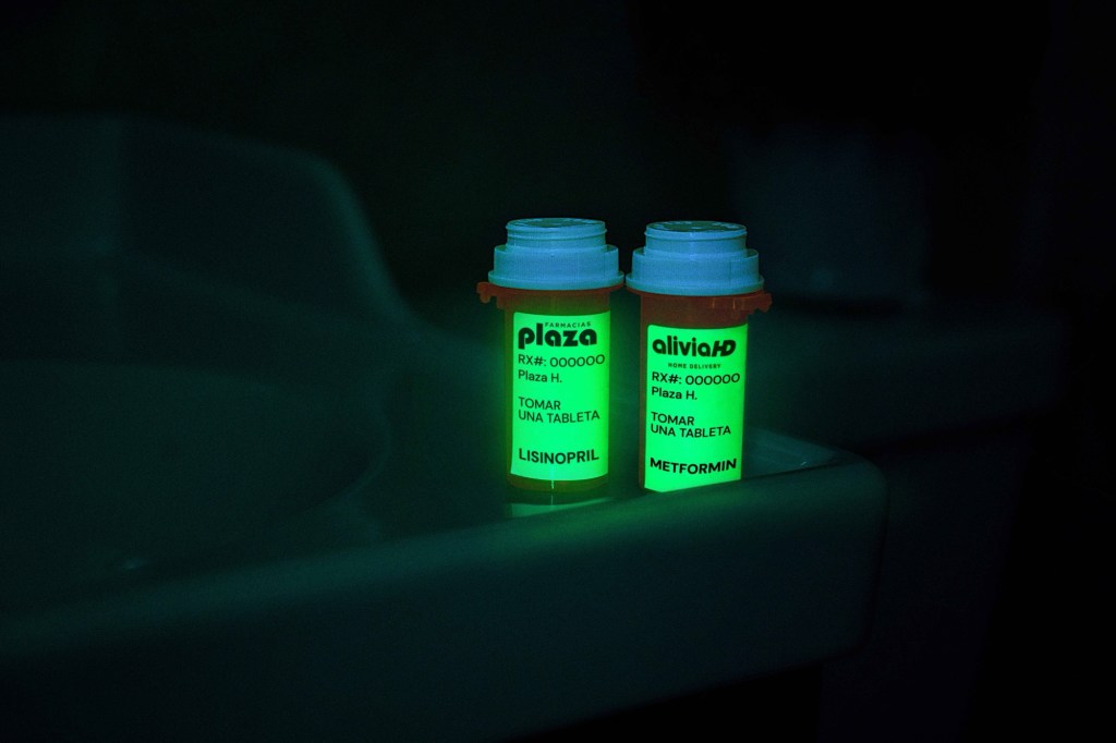 Dos envases de medicamentos con etiquetas fosforescentes brillan en la oscuridad sobre una superficie de baño. Uno de los envases es de Farmacias Plaza y el otro de Alivia Home Delivery. Ambos muestran información sobre el medicamento y las instrucciones de uso, permitiendo su lectura en ausencia de luz.