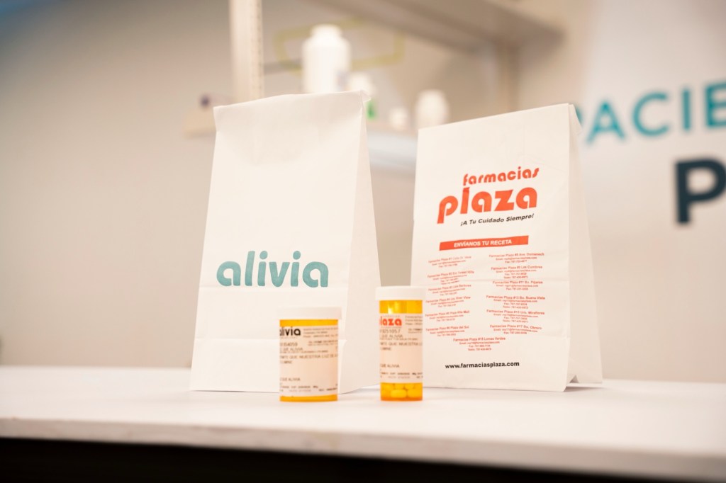 Dos bolsas de papel, una de Alivia y otra de Farmacias Plaza, se encuentran sobre un mostrador junto a dos envases de medicamentos con etiquetas estándar. El fondo muestra estantes con productos farmacéuticos en un entorno de farmacia.